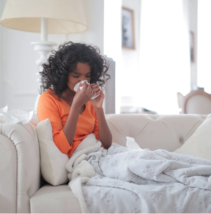 woman on couch blowing her nose. Everything in background (living room) is white. She's wearing an orange shirt.