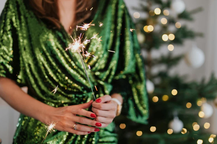 Woman in a green sequined dress holding a sparkling sparkler in front of a Christmas tree with warm holiday lights.