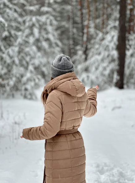 Woman standing outdoors in a snowy winter landscape wearing a long coat and knit hat, representing the need for extra skin care and protection during cold weather.