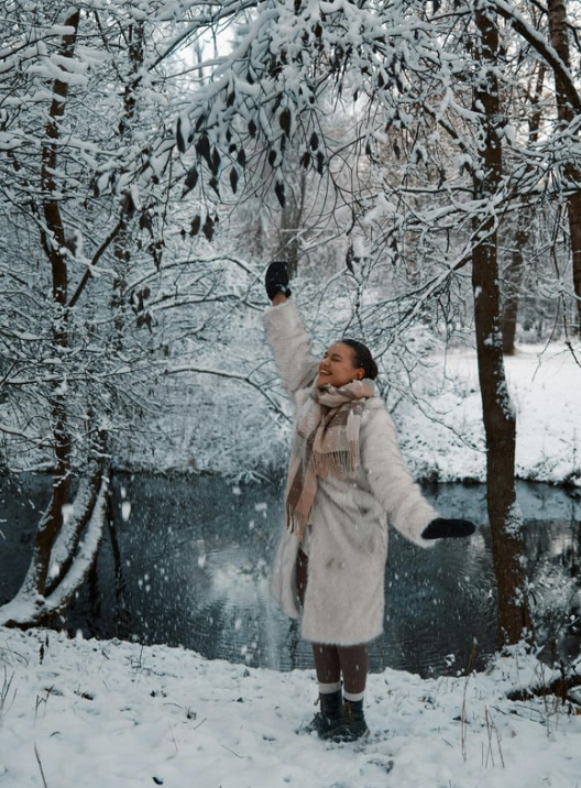 Woman standing outdoors in a snowy winter landscape, enjoying falling snow while dressed warmly, representing winter self-care and seasonal skin protection.