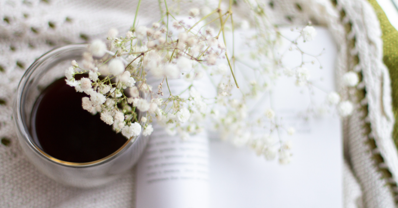 Soft wellness image featuring a cup of tea, delicate white flowers, and an open book on a cozy blanket, symbolizing emotional wellness, self-care, and finding peace during emotionally heavy times.